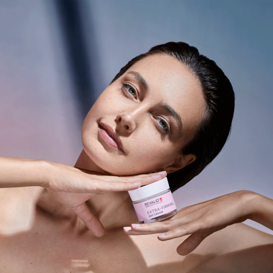 Woman holding a pink-labeled “Extra-Firming Day Cream” jar under her chin, gently framing her face with her hands.